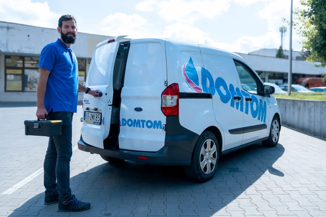 Serwisant DOMTOM z walizką narzędziową przy oznakowanym aucie serwisowym